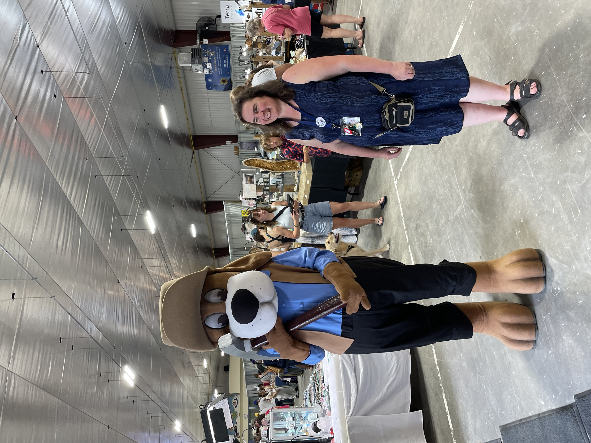 A woman in a panda costume headpiece walking inside a large indoor event space with booths, tables, and other attendees, possibly at a fair or market.