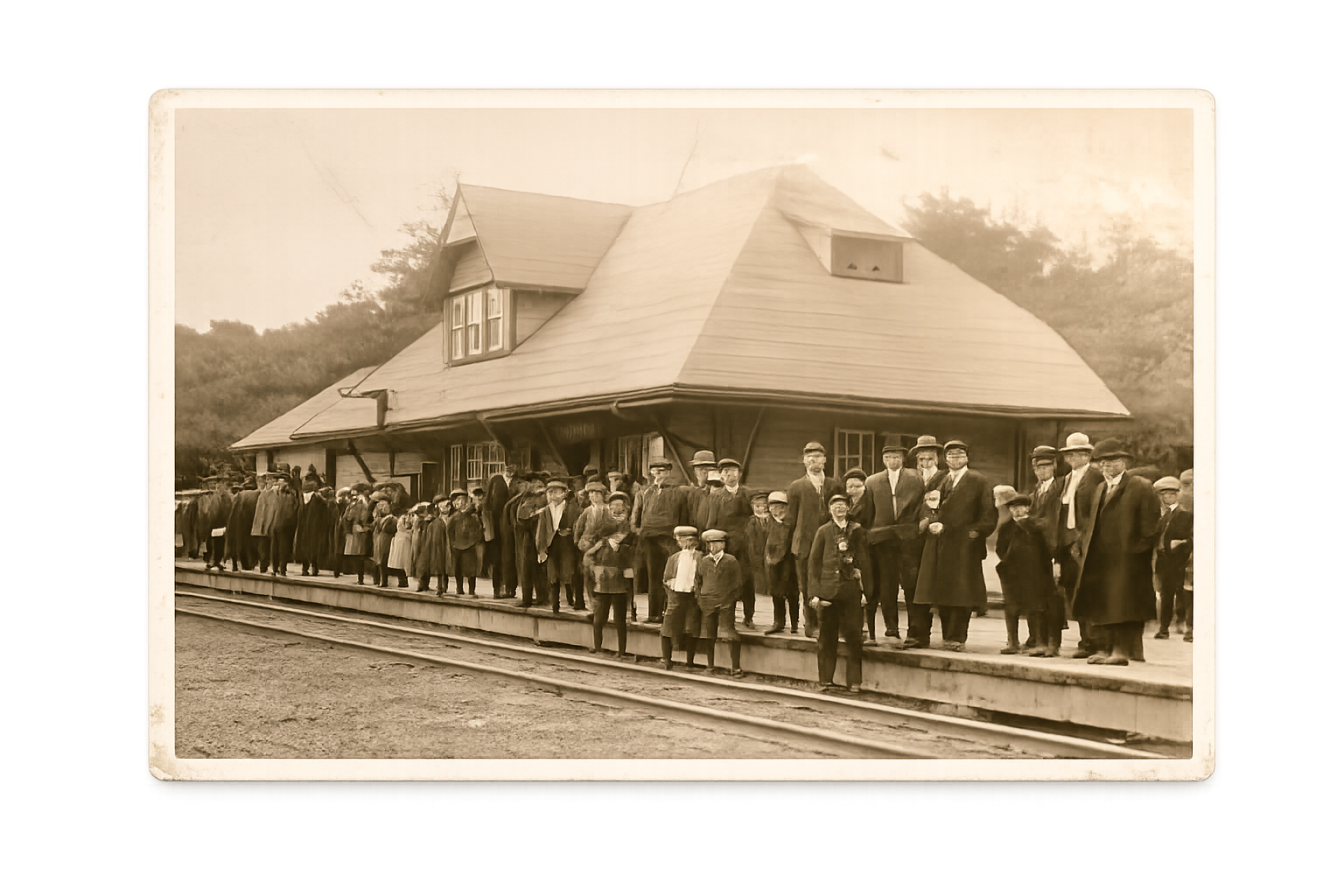 A line of people, including children, standing on a train platform in front of an old house with a steep roof and dormer windows.