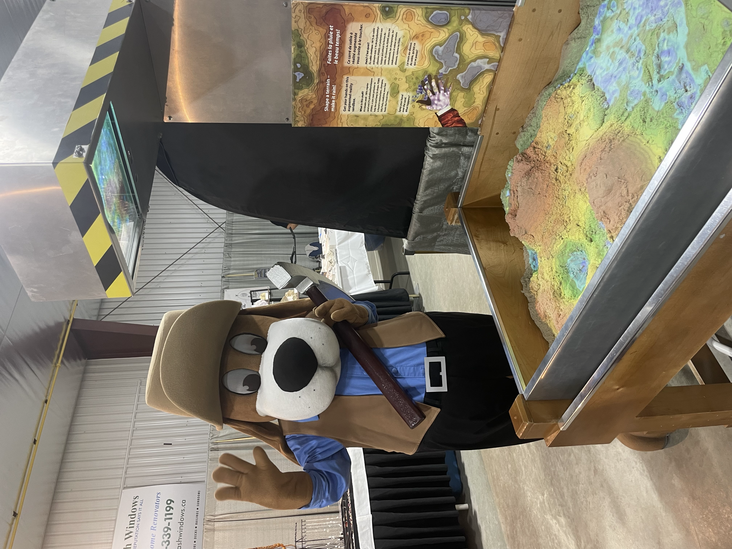 A person dressed in a hooded costume with large eyes and ears, resembling a cute animal, stands behind a colorful topographic map on a table. The person is making a hand gesture, and there is informational signage behind them at an indoor event or exhibition.