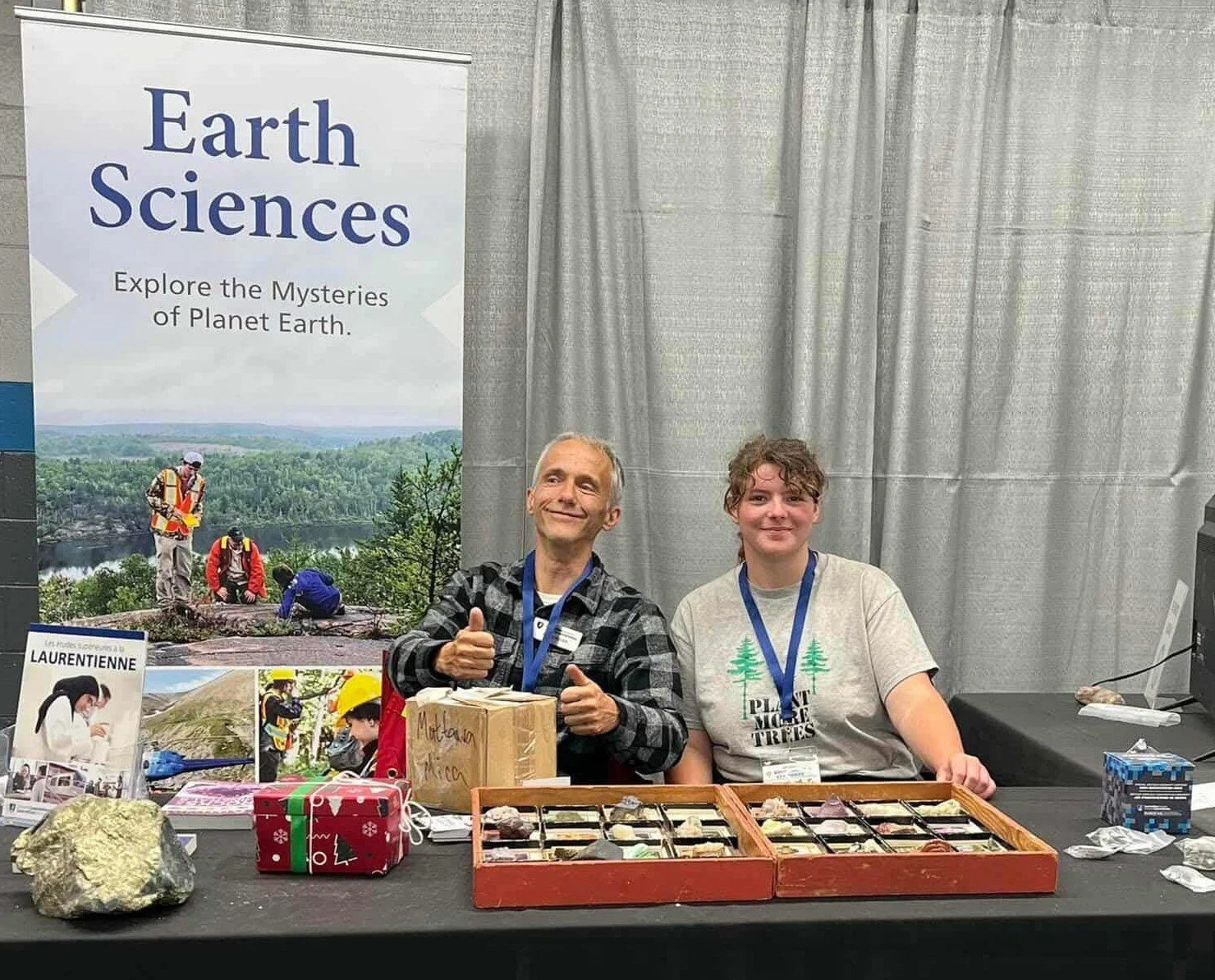 Two people sitting at a table during an Earth sciences event. The person on the left is giving a thumbs-up and smiling, while the person on the right is smiling. The table displays rocks, minerals, and informational materials. A large poster behind t