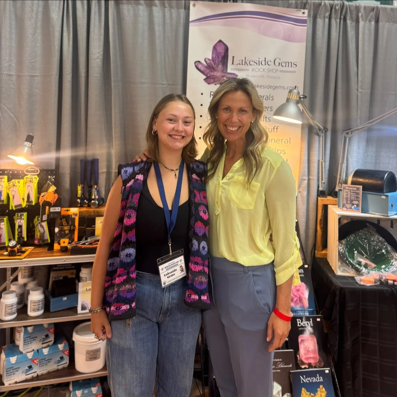 Two women standing together at a booth, smiling for the camera, with jewelry and gemstone products on display behind them.
