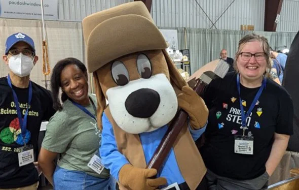 Four people in a group photo, three smiling women and a person in a dog costume with a beige hat, blue shirt, and holding a stick. One woman wears a face mask and the others have lanyards with badges. They are indoors at an event with industrial wall