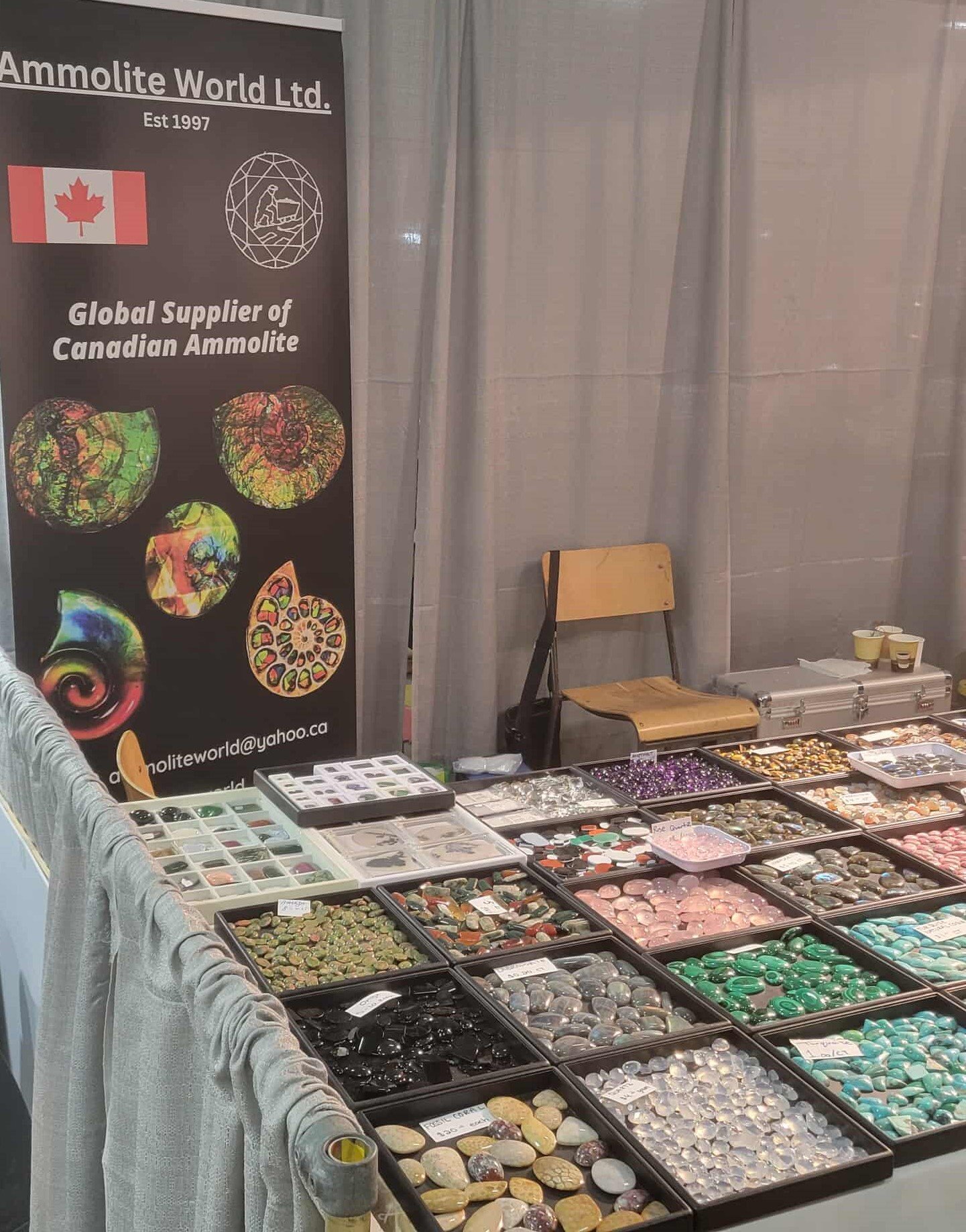 A booth at a market displays various polished stones and gemstones arranged in black trays. A banner nearby reads 'Ammolite World Ltd.' and features a Canadian flag, with text indicating it is a supplier of Canadian ammolite. There is a wooden chair 