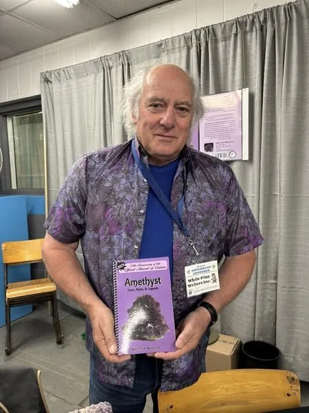 An older man with white hair holding a book titled "Amethyst" at a book fair or event.