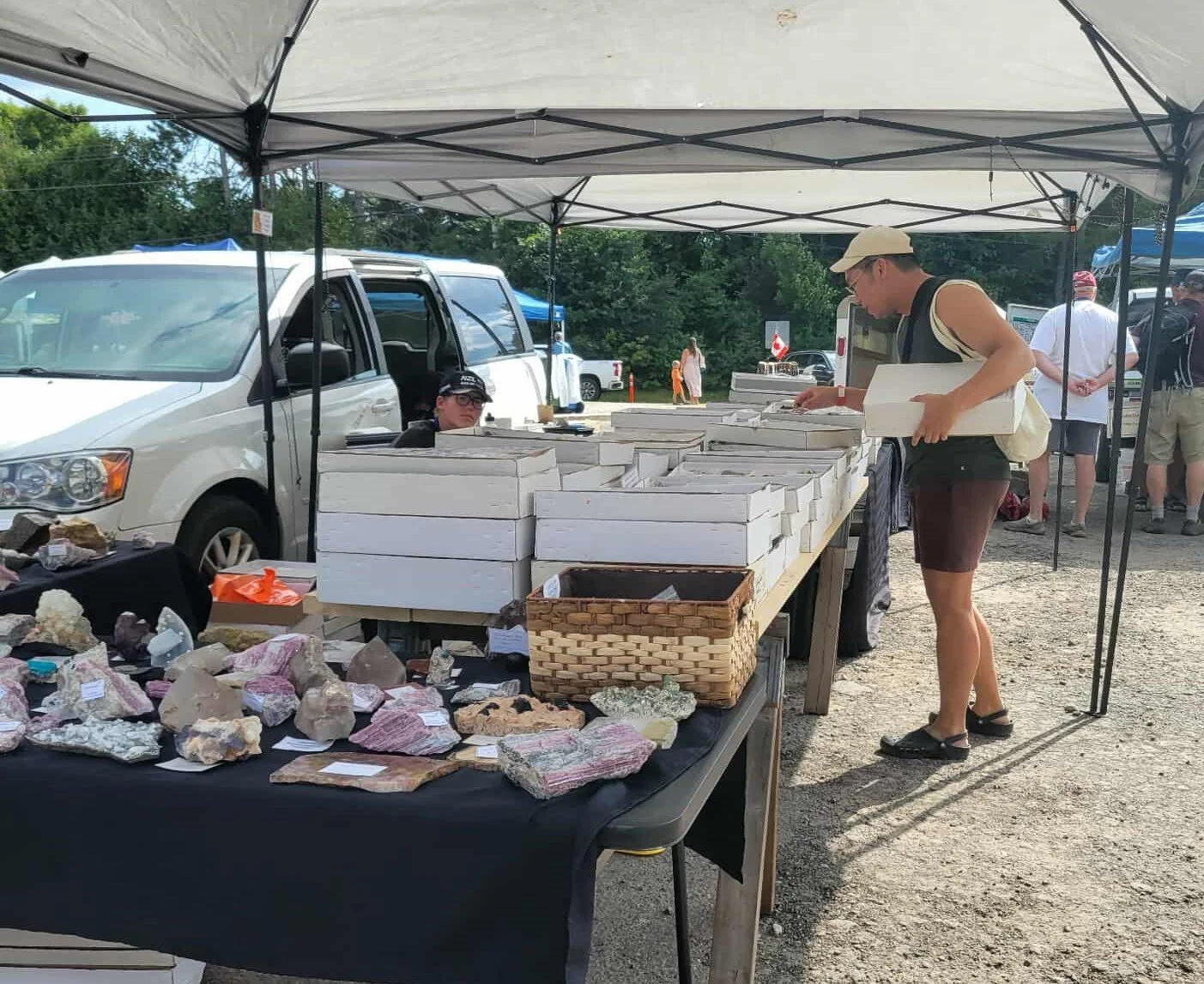 A woman browsing gemstones at an outdoor market stall under a canopy, with rocks and minerals displayed on a black table in front of her. Several other people and vehicles are visible in the background.