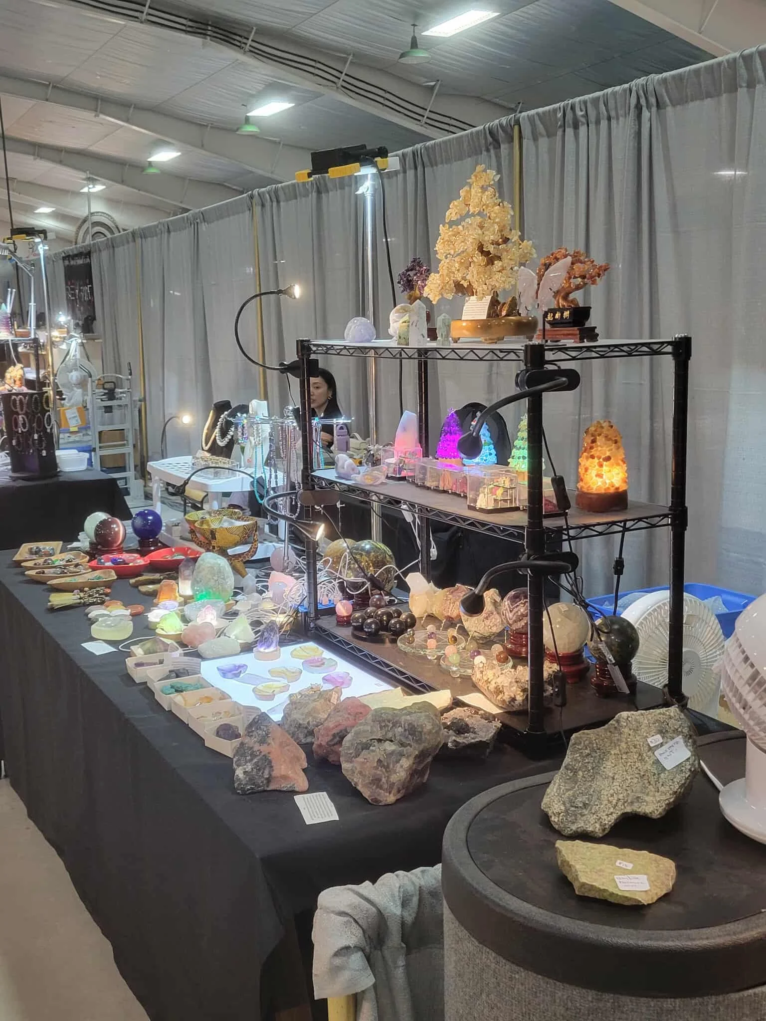 An indoor gemstone and mineral exhibit with various rocks, crystals, and mineral specimens displayed on tables and shelves. There are decorative mineral lamps and illuminated displays, with a curtain backdrop and a person behind the booth.