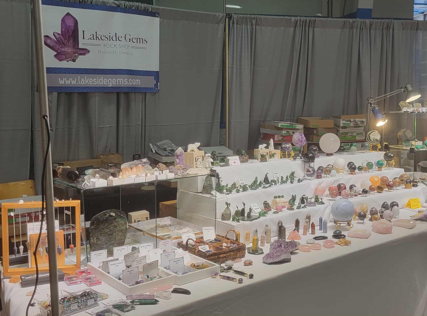 Jewelry and decorative glass stones displayed on tables at a gem and mineral show, with a banner reading "Lakeside Gems ROCK SHOP" in the background.