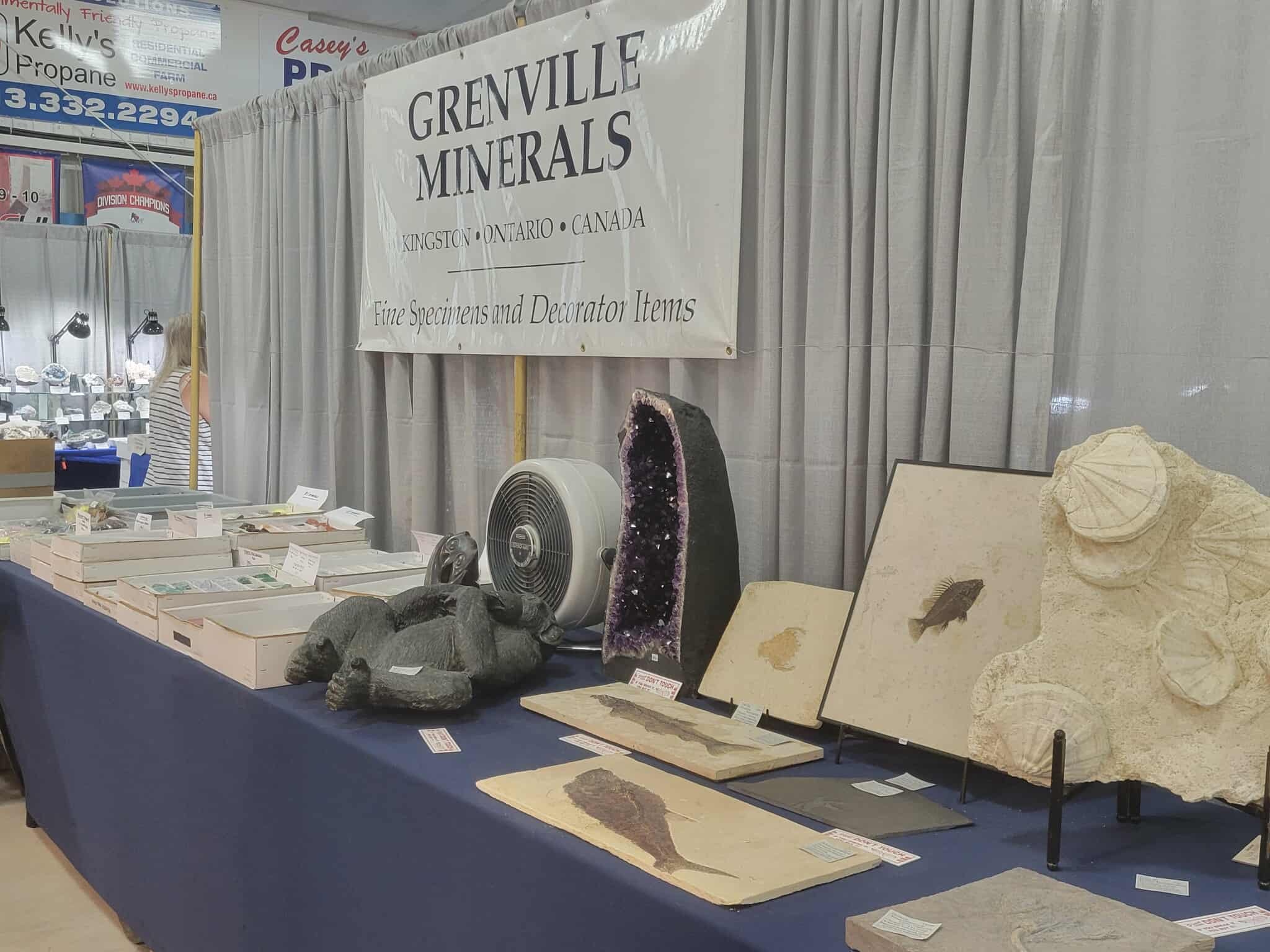 Display booth at a mineral show featuring a large banner that reads 'Grenville Minerals, Kingston Ontario Canada, Fine Specimens and Decorator Items.' The table holds various mineral specimens, rocks, and decorative stone items, including an amethyst