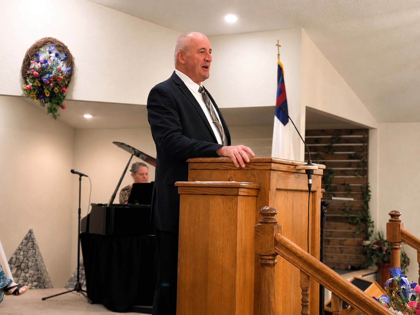 A man in a black suit and tie standing at a wooden podium speaking, with a woman playing a piano in the background, and a flag with red, blue, and white colors and a cross in the background.