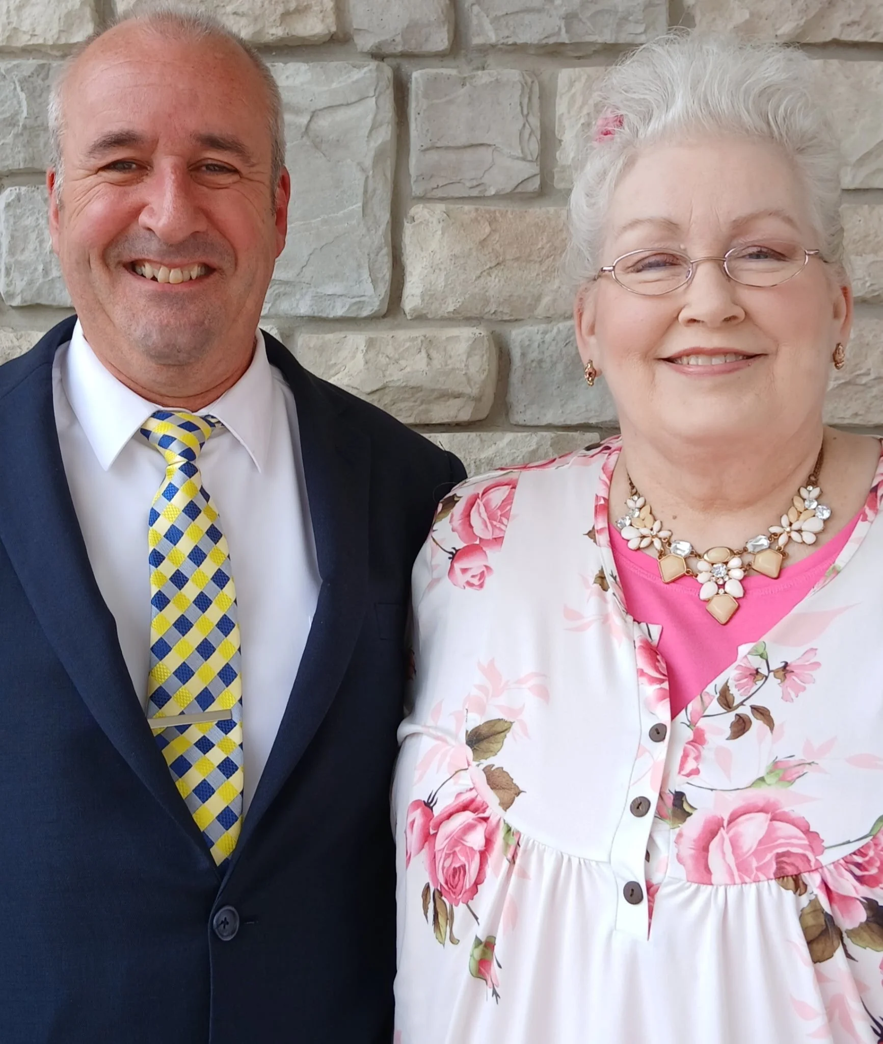 A smiling man wearing a dark suit, white shirt, and a yellow and blue patterned tie, standing next to a smiling woman with white hair, glasses, floral earrings, and a floral dress. They are against a stone wall background.