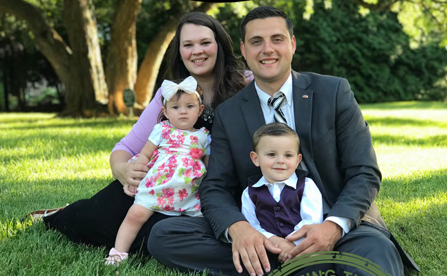 Family of four sitting on grass in a park, smiling together, with trees and sunlight in the background.