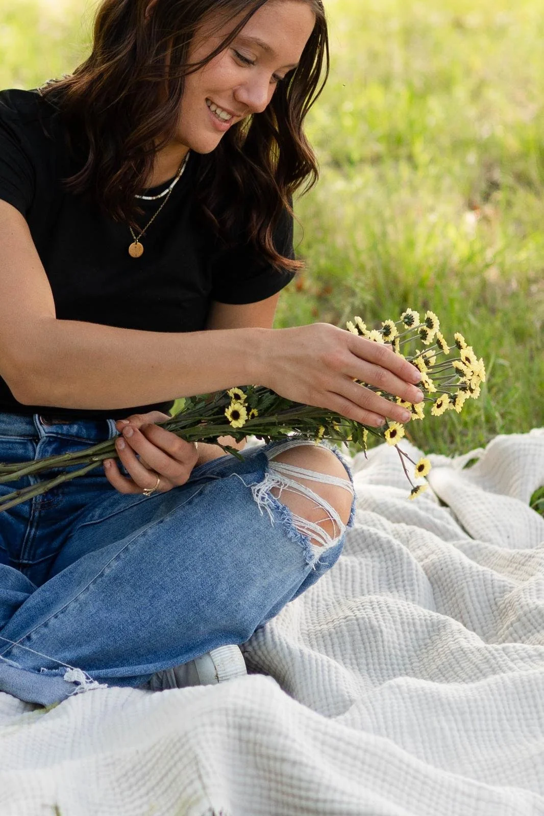 A woman with brown hair, wearing a black t-shirt and ripped jeans, sitting outdoors on a cream-colored blanket in a grassy field, holding a bouquet of yellow and black daisies, smiling and looking at the flowers.