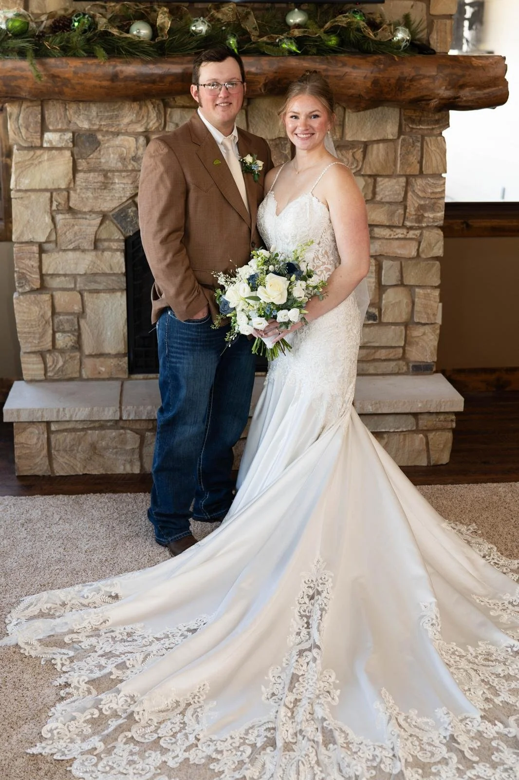 A bride and groom standing in front of a stone fireplace decorated with greenery and silver ornaments, smiling for a wedding photo. The bride is wearing a lace wedding gown and holding a bouquet of white and dark blue flowers. The groom is dressed in
