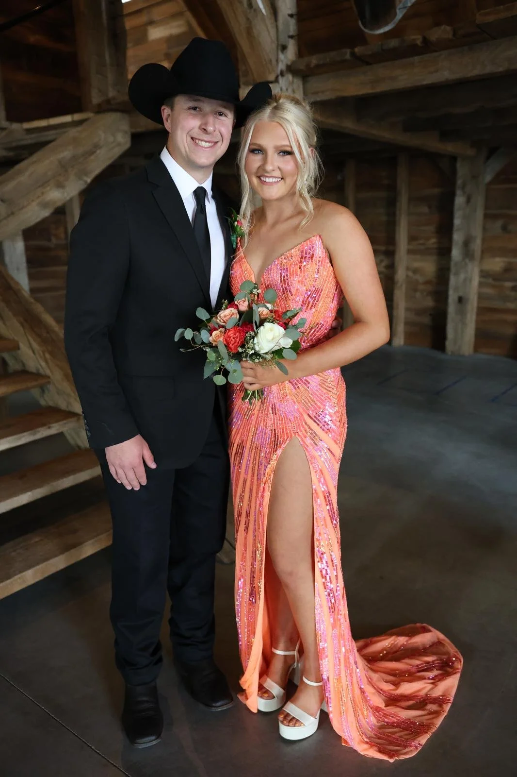 A young man in a black suit and cowboy hat and a young woman in a peach-sequined dress holding a bouquet of flowers pose together inside a rustic wooden barn.