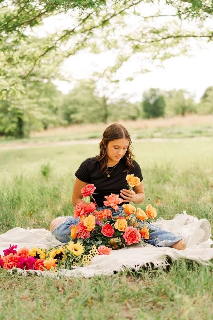 A young woman is sitting on a white blanket outdoors in a grassy field, arranging a colorful bouquet of flowers with pink, orange, and yellow blooms surrounded by trees.
