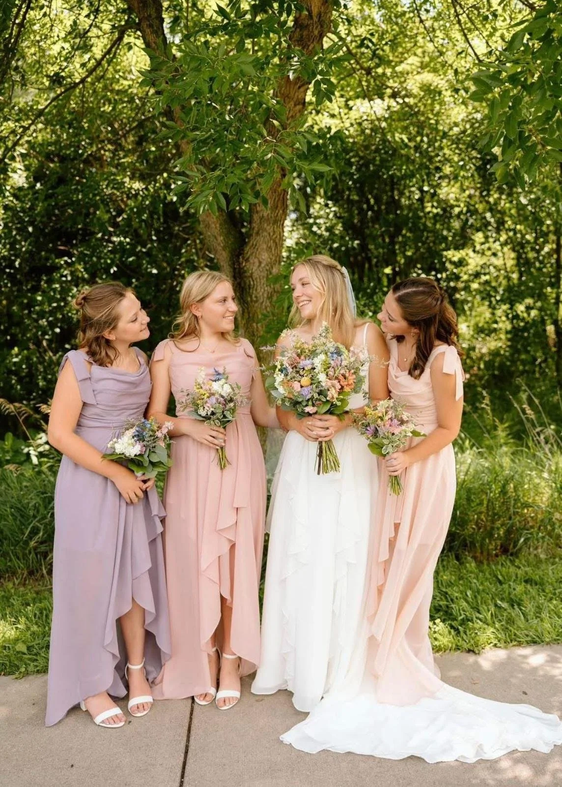 Four women in pastel dresses holding bouquets, standing outdoors under a large tree, smiling and looking at each other.