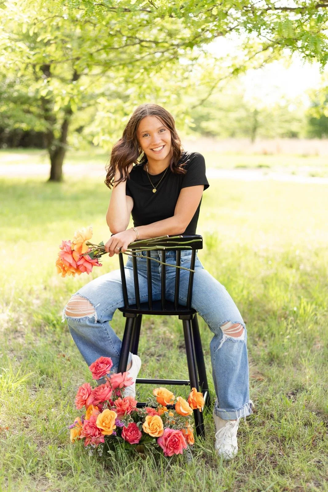 A young woman with brown hair sitting on a black chair outdoors, holding a bouquet of pink and orange flowers, with more flowers on the ground in front of her. She is smiling, wearing a black t-shirt, ripped jeans, and white sneakers, in a sunny park with green trees and grass.