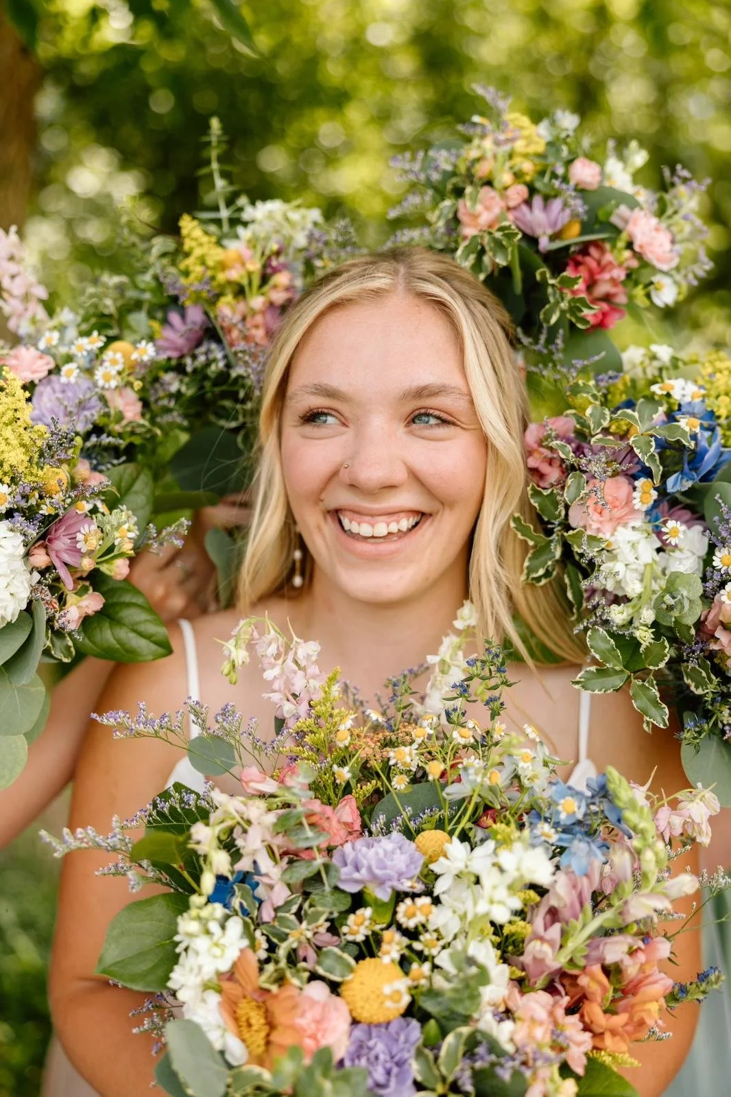 A young woman with blonde hair and a nose piercing smiling in an outdoor setting, surrounded by colorful flowers in a floral wreath and bouquet.