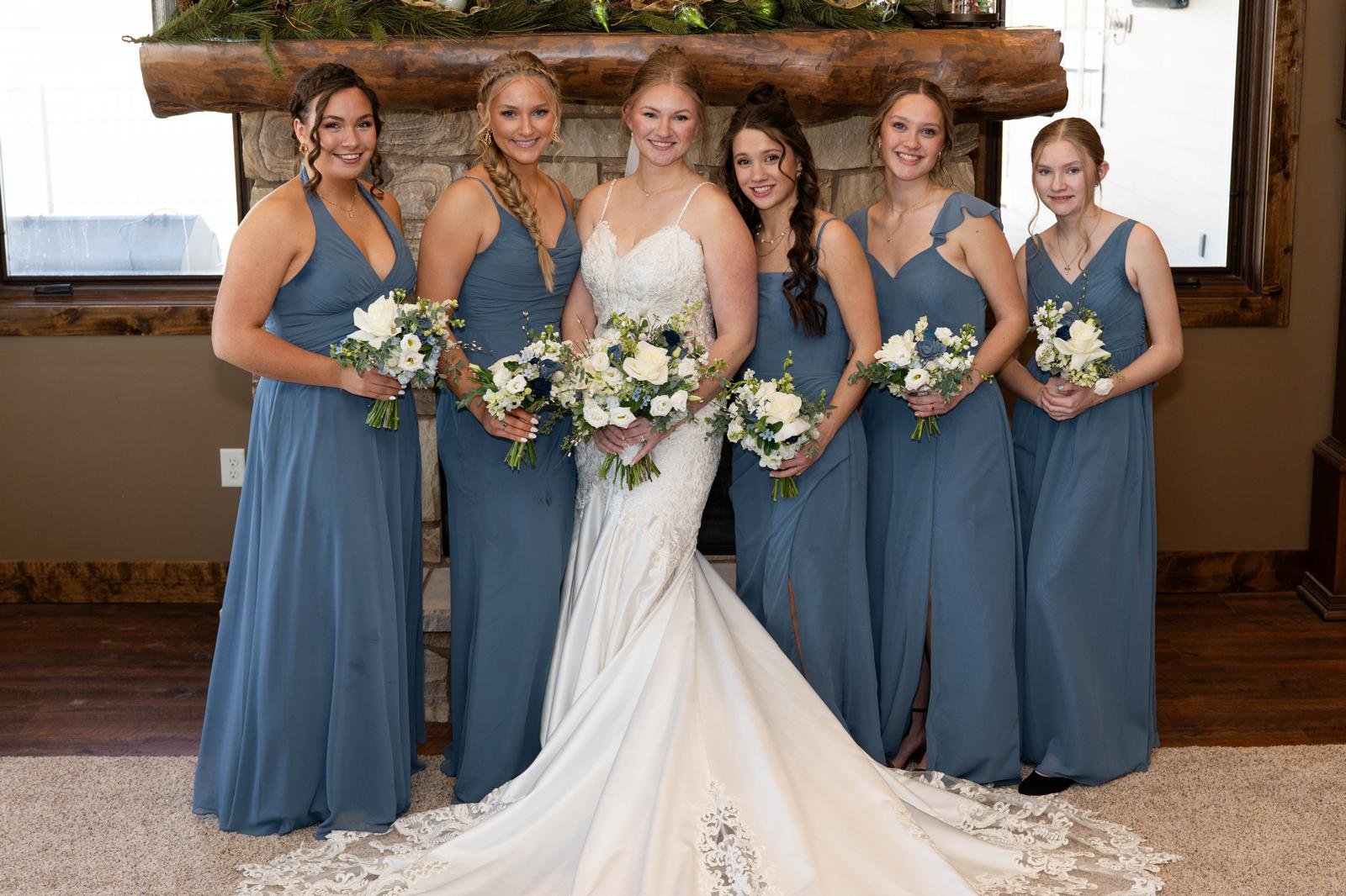 A bride in a white wedding gown standing with six bridesmaids in matching blue dresses, holding bouquets of white and blue flowers, in front of a stone fireplace with a wooden mantel.