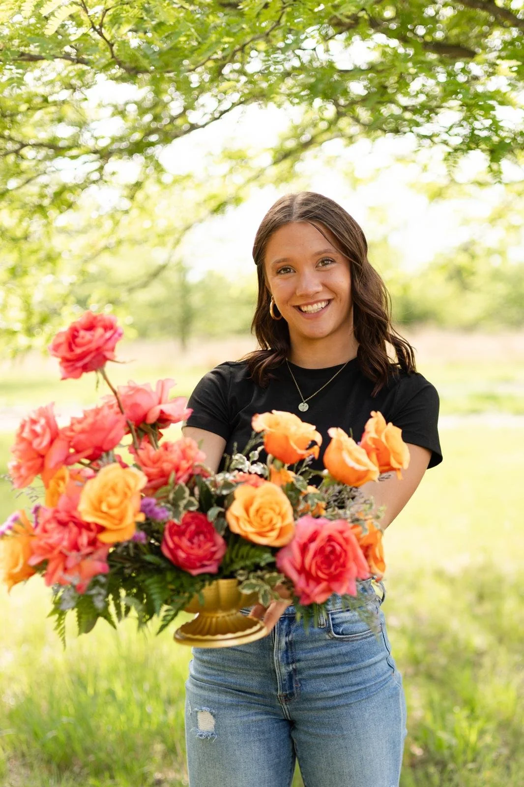 A young woman holding a large bouquet of colorful flowers outdoors, smiling at the camera, with green trees and sunlight in the background.