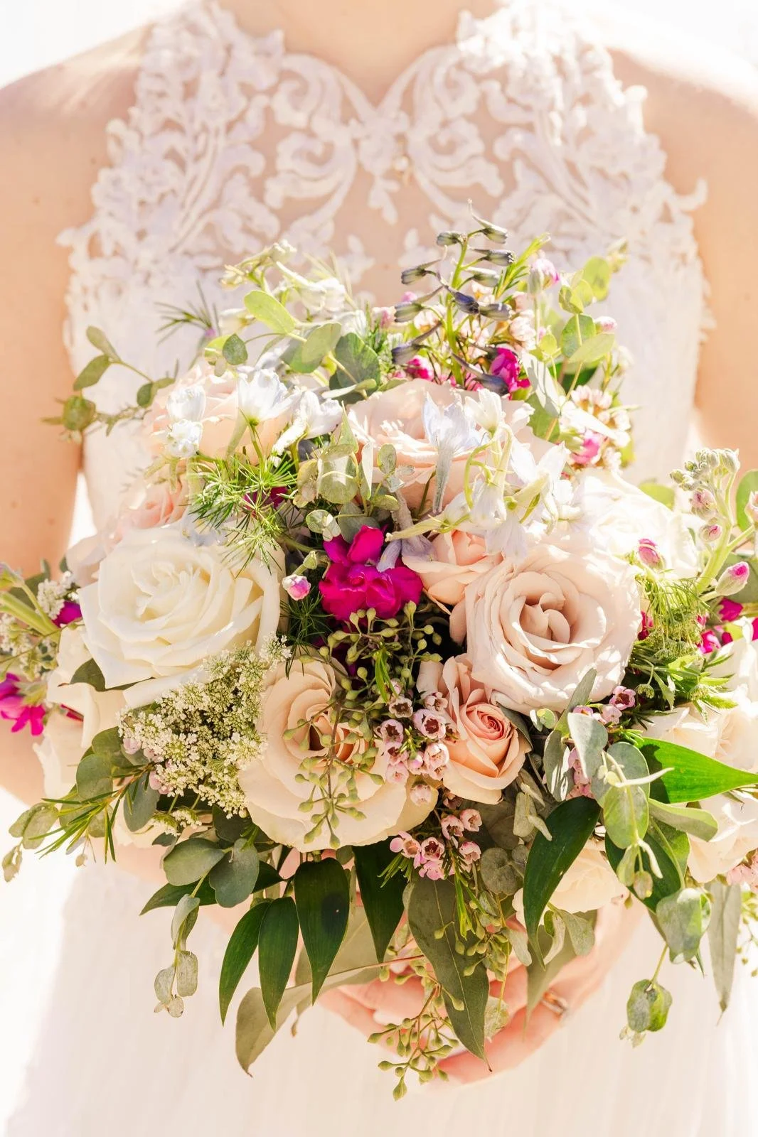 Close-up of a bride holding a bouquet of white, pink, and purple flowers, with a lace wedding dress in the background.