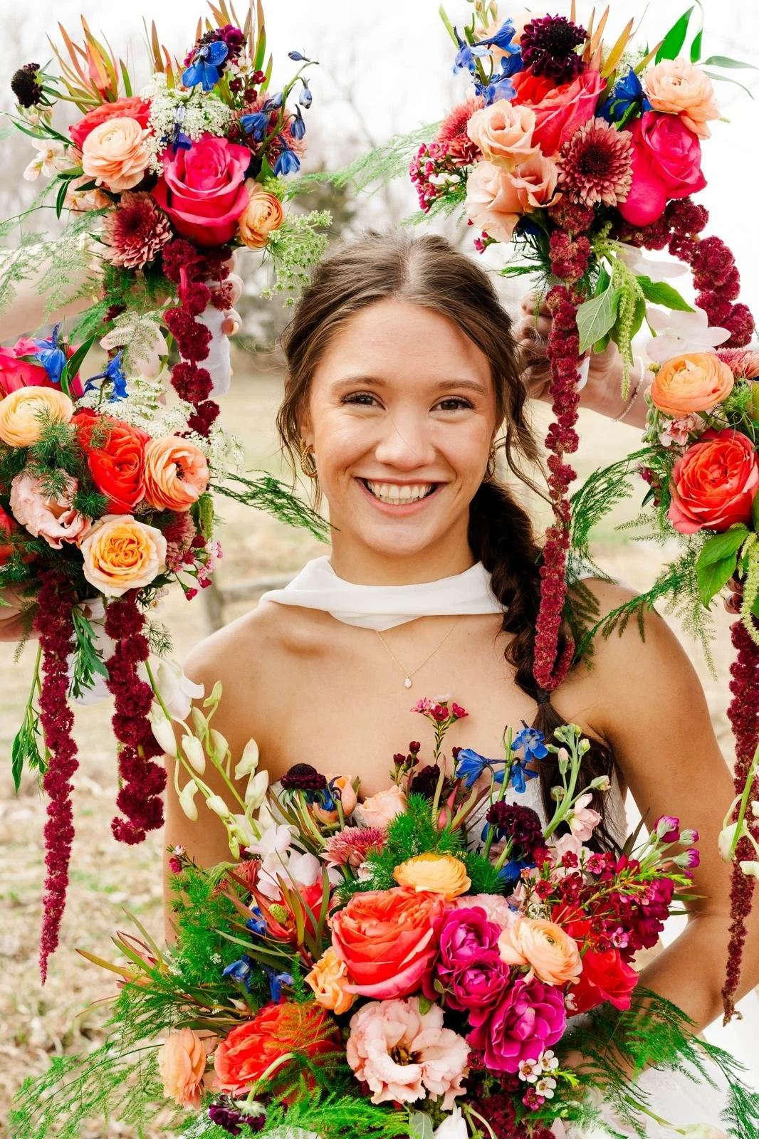 A smiling young woman in a white dress is holding a large floral wreath with colorful flowers surrounding her face. She has her hair in a side braid and is outdoors with blurred trees in the background.