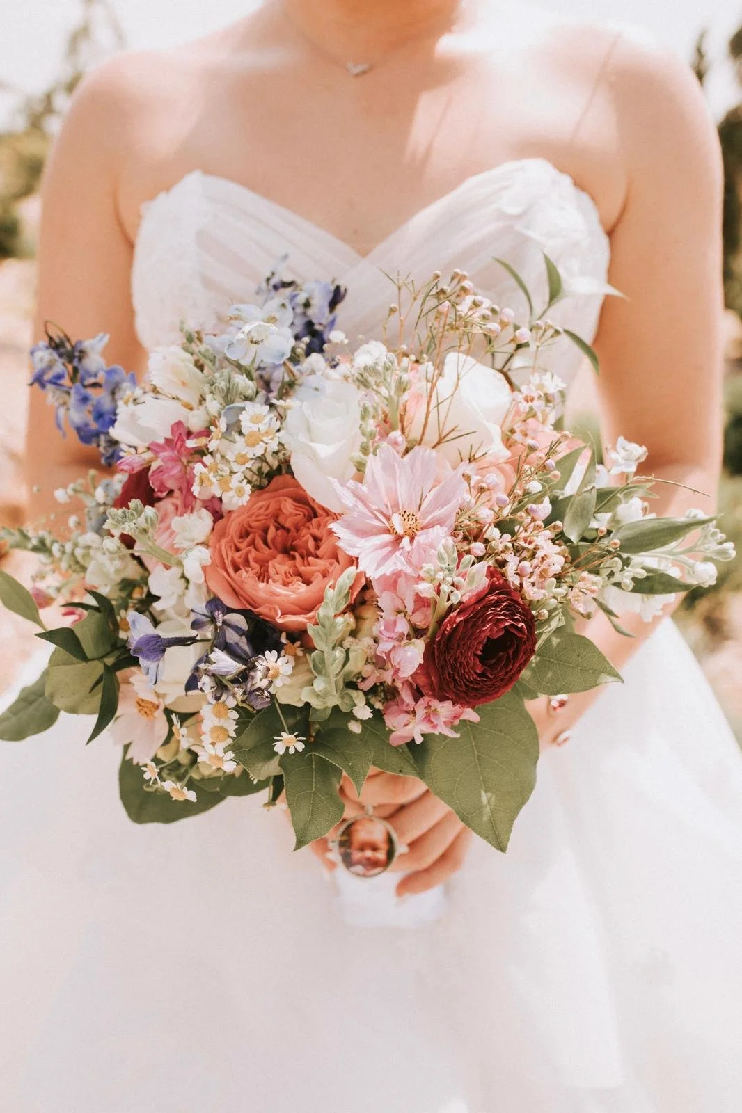 Bride holding a bouquet of colorful flowers, including pink, red, and white roses, with green leaves, against a blurred outdoor background.