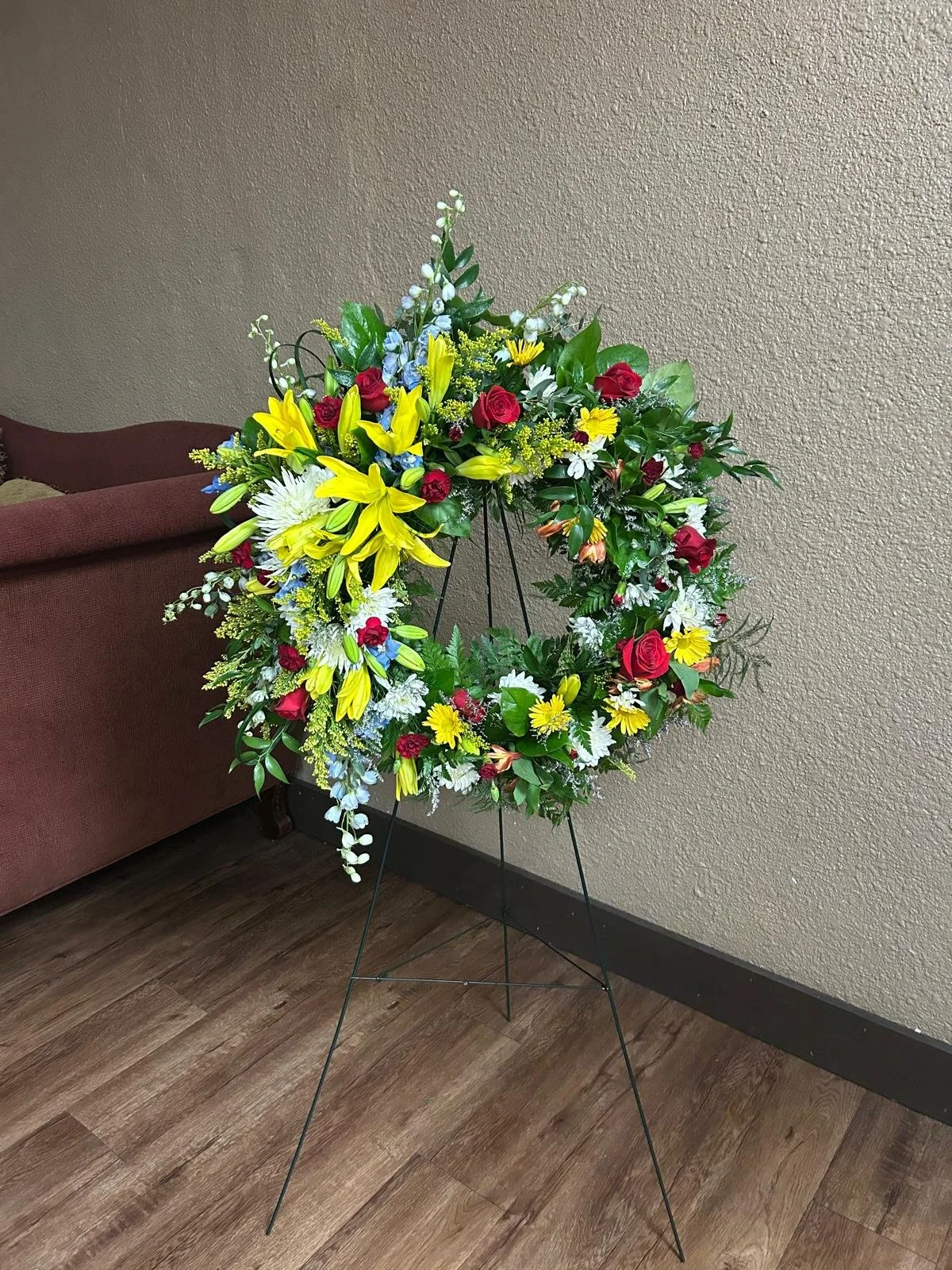 Lg Hanging Wreath ft yellow lilies, red roses and light blue delphinium. 