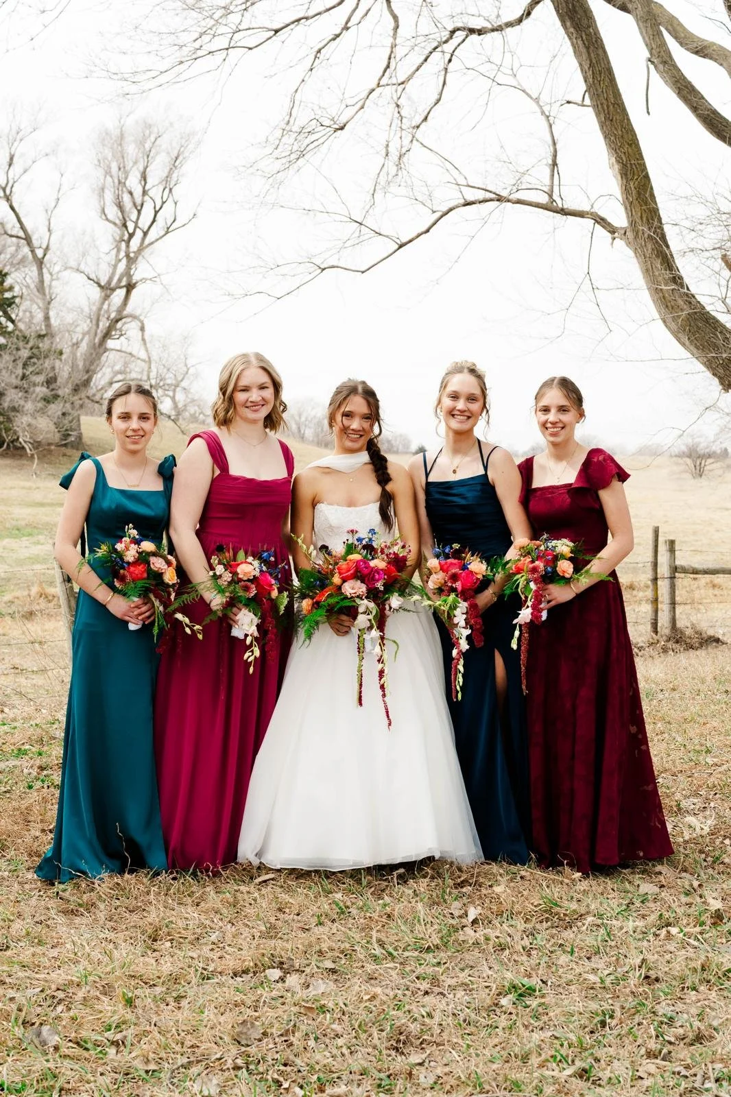 Group of five women in colorful dresses standing outdoors in a field, holding bouquets of flowers, during daytime.