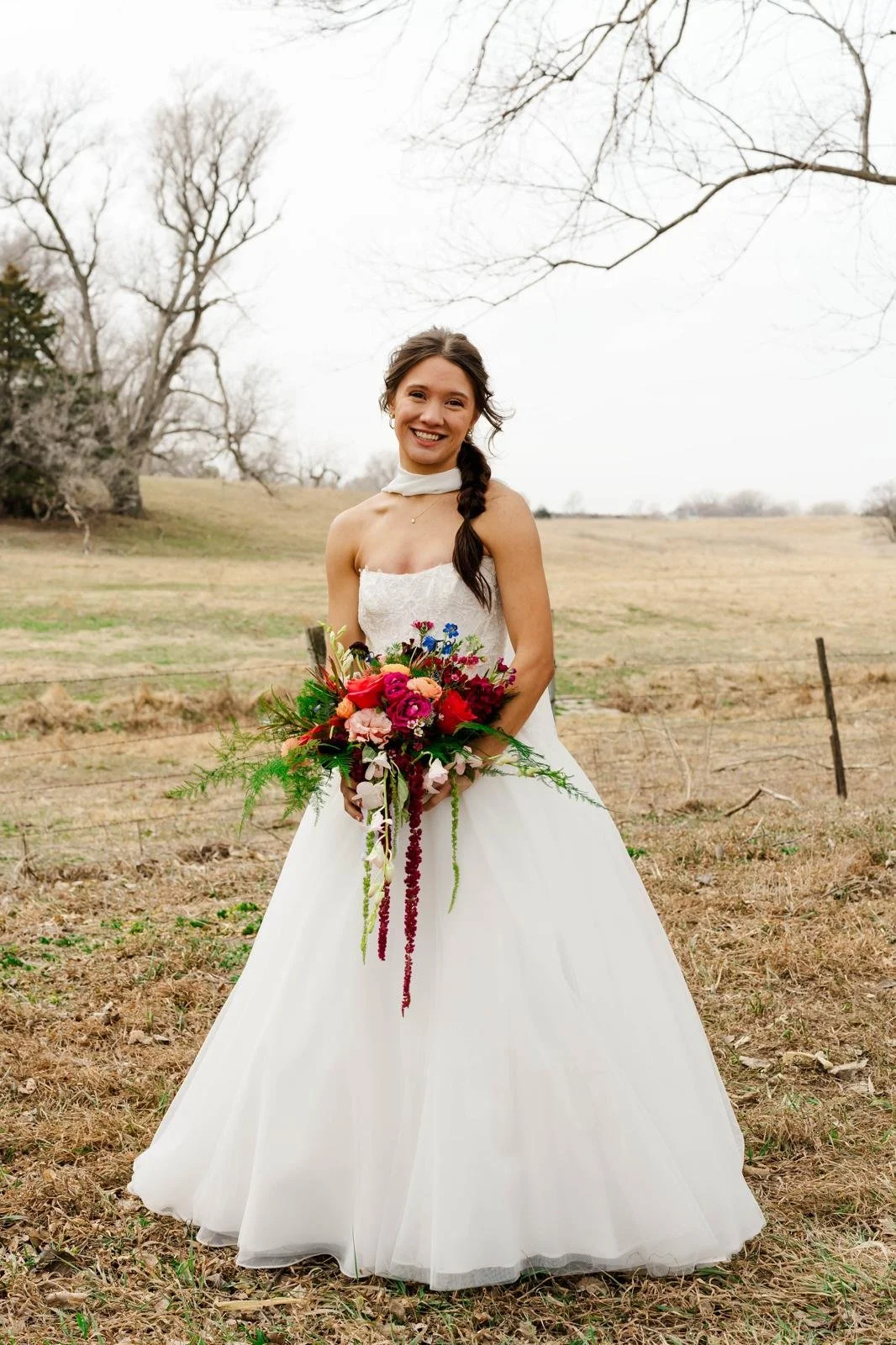 A smiling bride in a white wedding dress holding a vibrant bouquet of pink, red, purple, and white flowers standing outdoors in a field with leafless trees in the background.