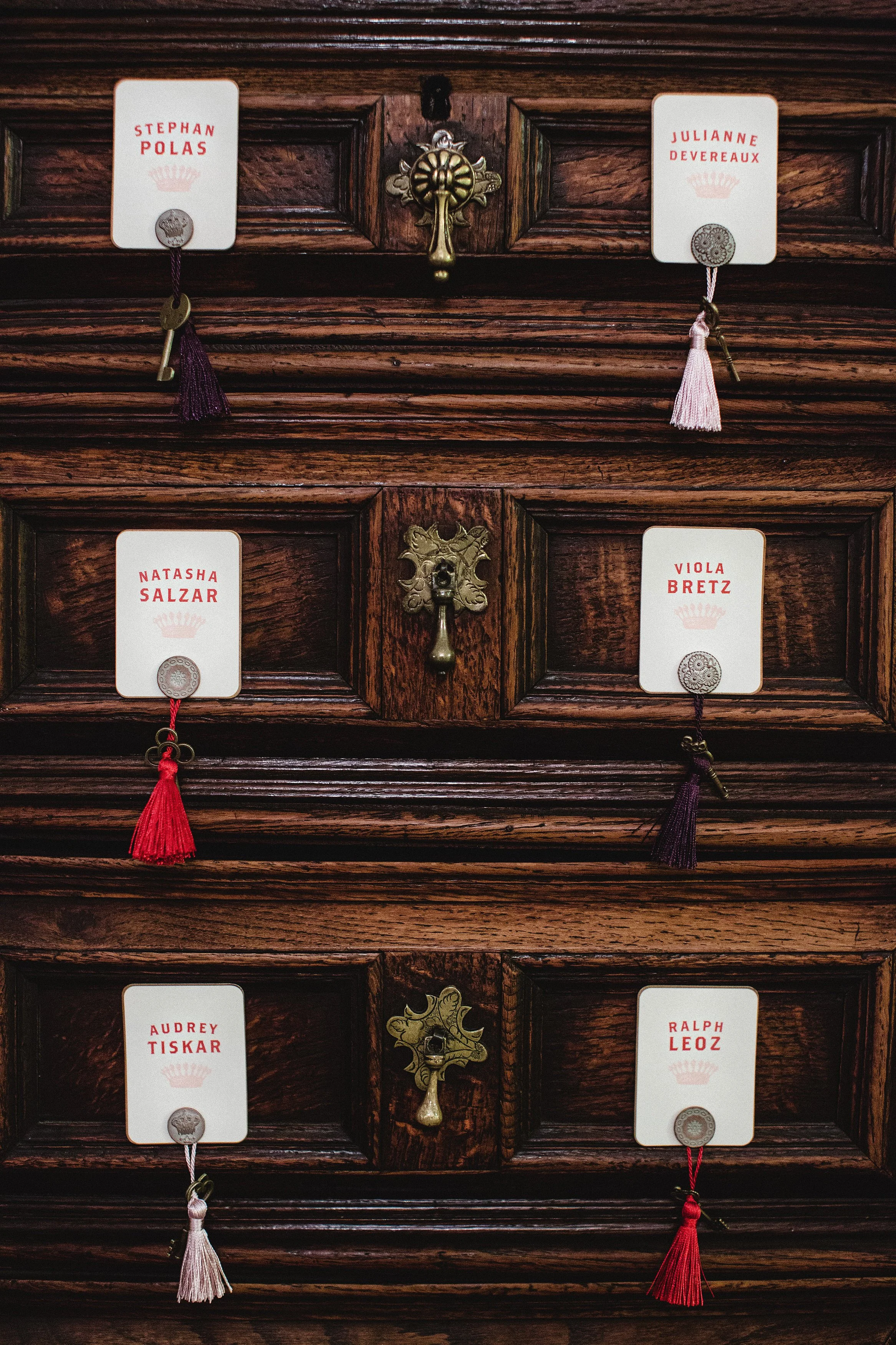 A wooden cabinet with small drawers, each labeled with name tags for characters like Stephan Polas, Julianne Devereaux, Natasha Salzar, Viola Bretz, Audrey Tiskar, and Ralph Leoz, with decorative tassels hanging from each label.