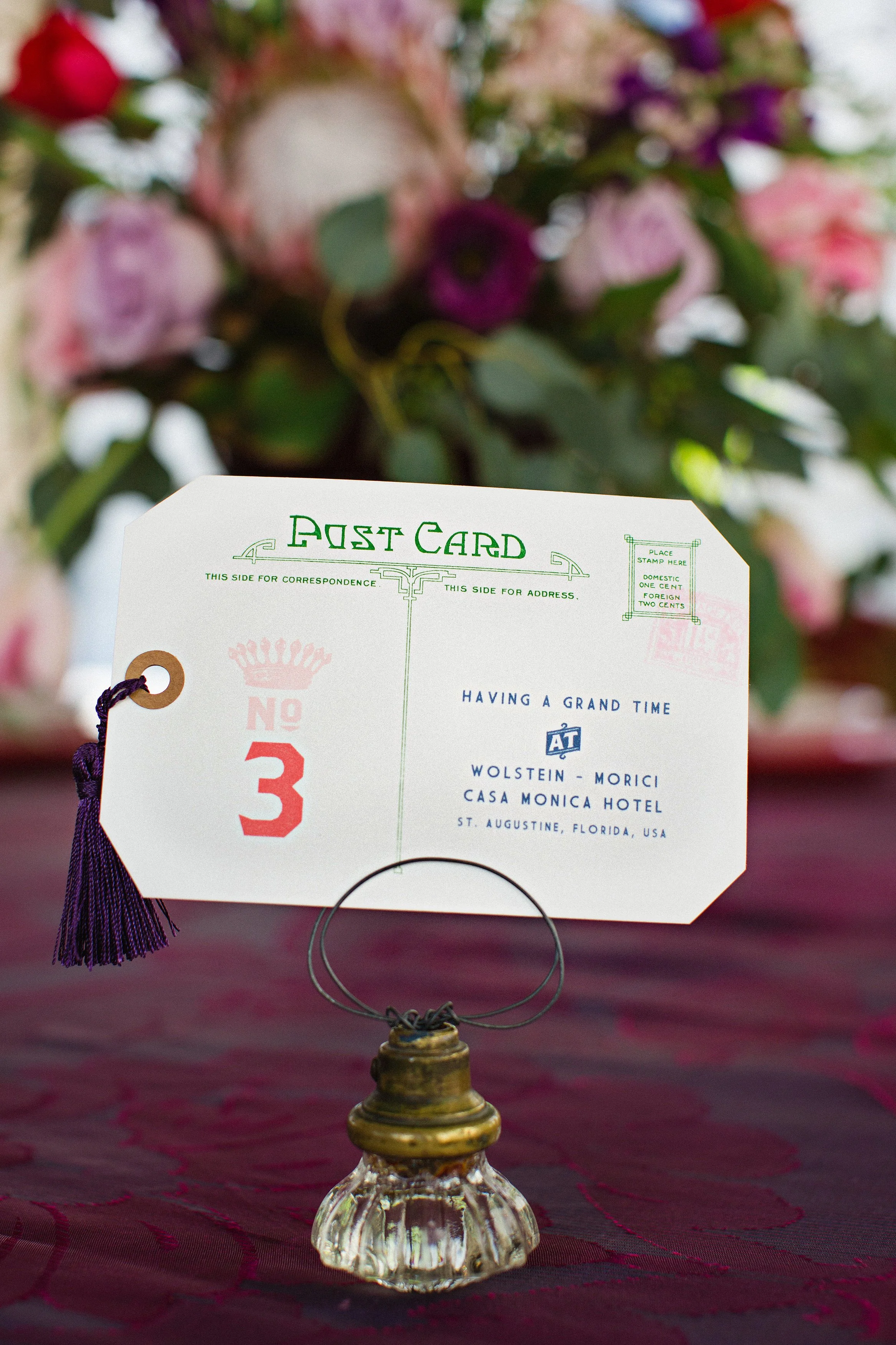 A vintage style postcard or table number card on a small glass stand, with a purple tassel on a string attached, placed on a table with a dark red tablecloth. In the blurred background, there is a flower arrangement with pink, purple, and white flowe