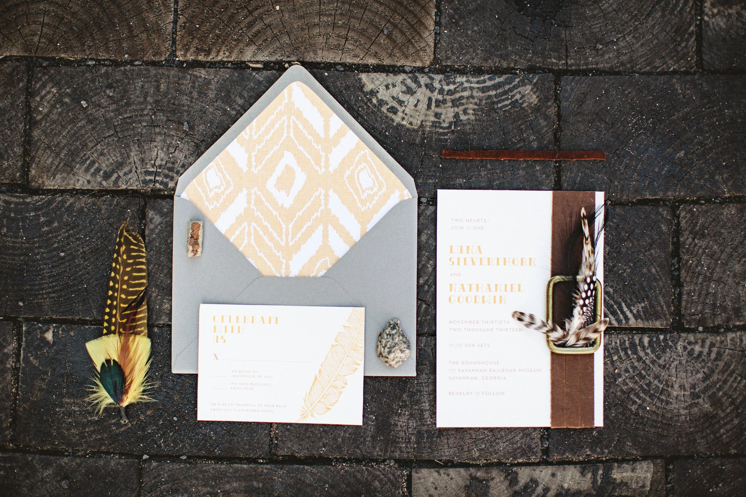 Wedding invitation suite with RSVP card, envelope, and decorative feathers and rocks, placed on a dark wooden surface.