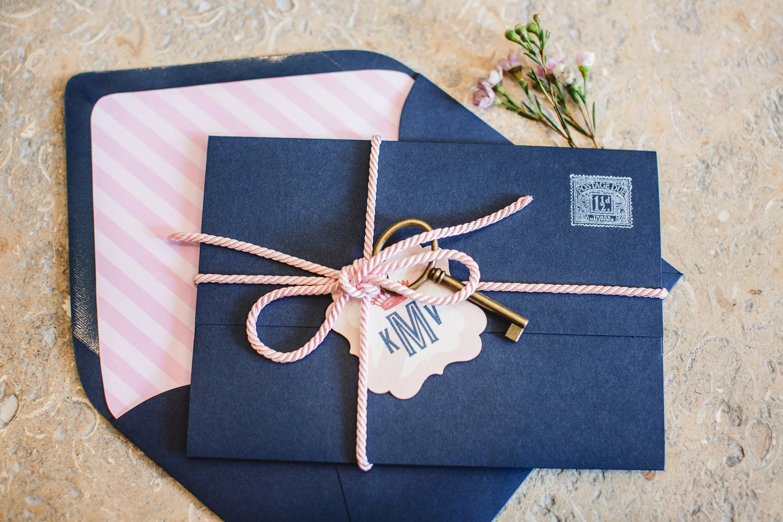 A navy blue envelope tied with pink and white striped string, with a vintage key, a small label marked "MAMY," and a sprig of pink flowers placed on a beige surface.