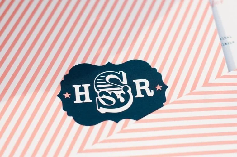Close-up of a black sticker with the letters H, S, and R, placed on a pink and white striped surface.