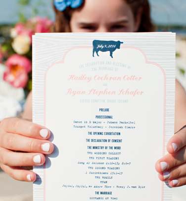 A young girl holding a white wedding invitation card with pink and blue text and a blue ribbon, outdoors with flowers in the background.