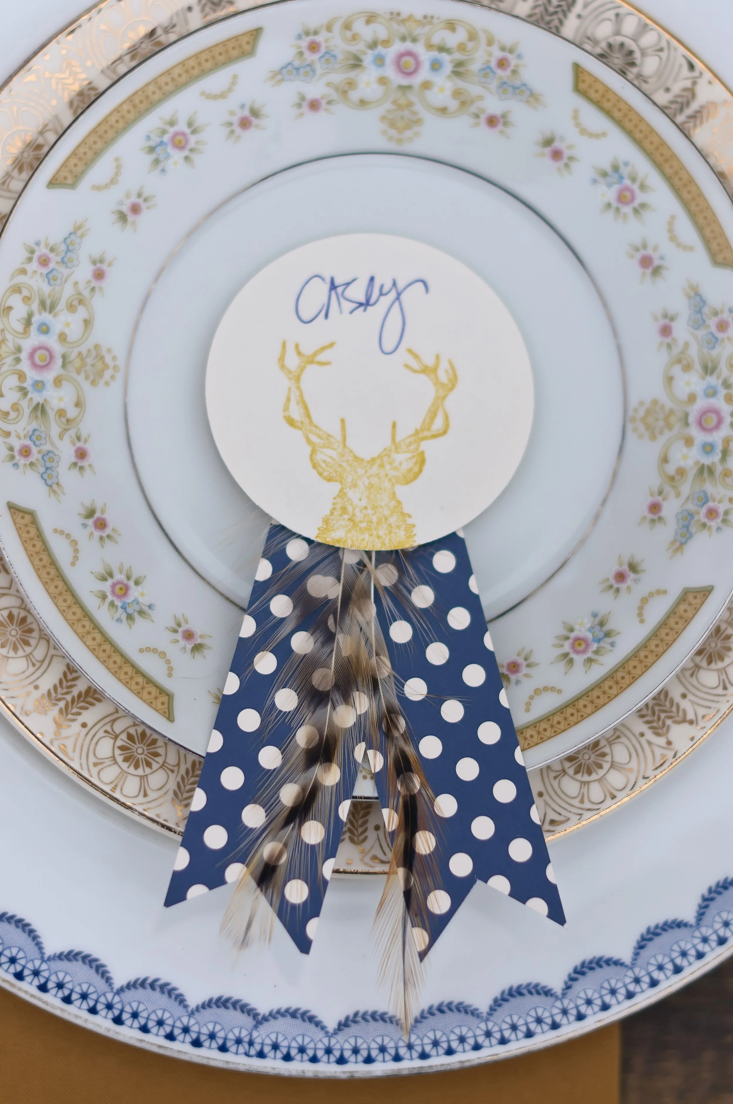 Decorative place setting with floral patterned china plates, a paper tag with a golden deer head illustration and a handwritten name, and a navy blue and white polka dot ribbon with feathers.