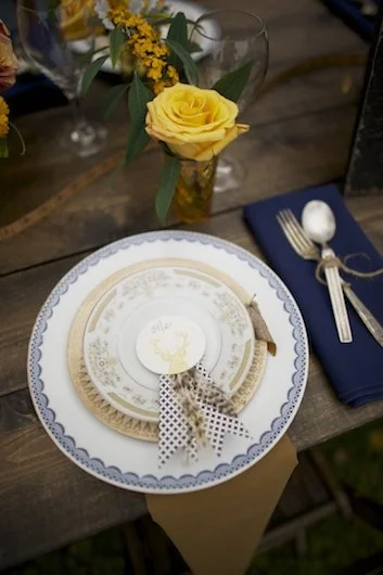 Plate setting with a yellow rose in a vase, napkin with gold utensils, and a small decorative item on a wooden table.