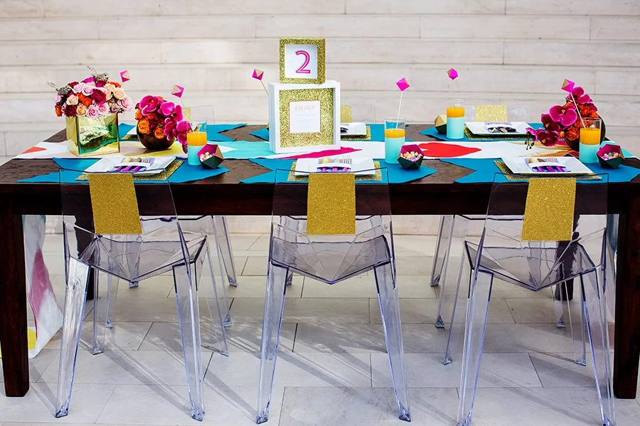 Decorated table for a celebration with floral centerpieces, colorful drinks, legal pad and pens, and a numbered sign, in front of a white wooden wall.