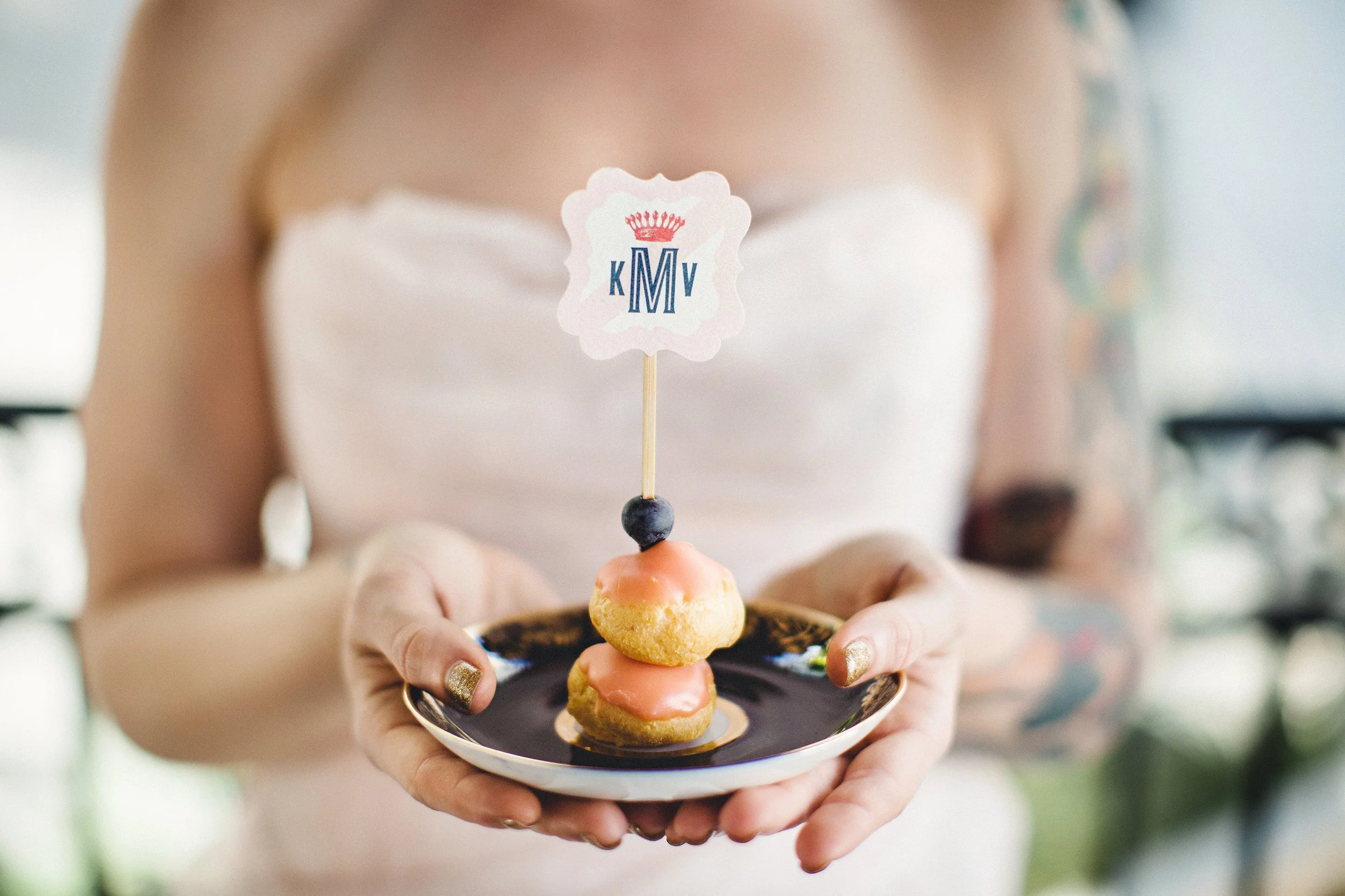 Person in a white strapless dress holding a black and gold plate with three small cream puffs topped with icing, with a small sign on a stick featuring a crown and initials 'K M V'.