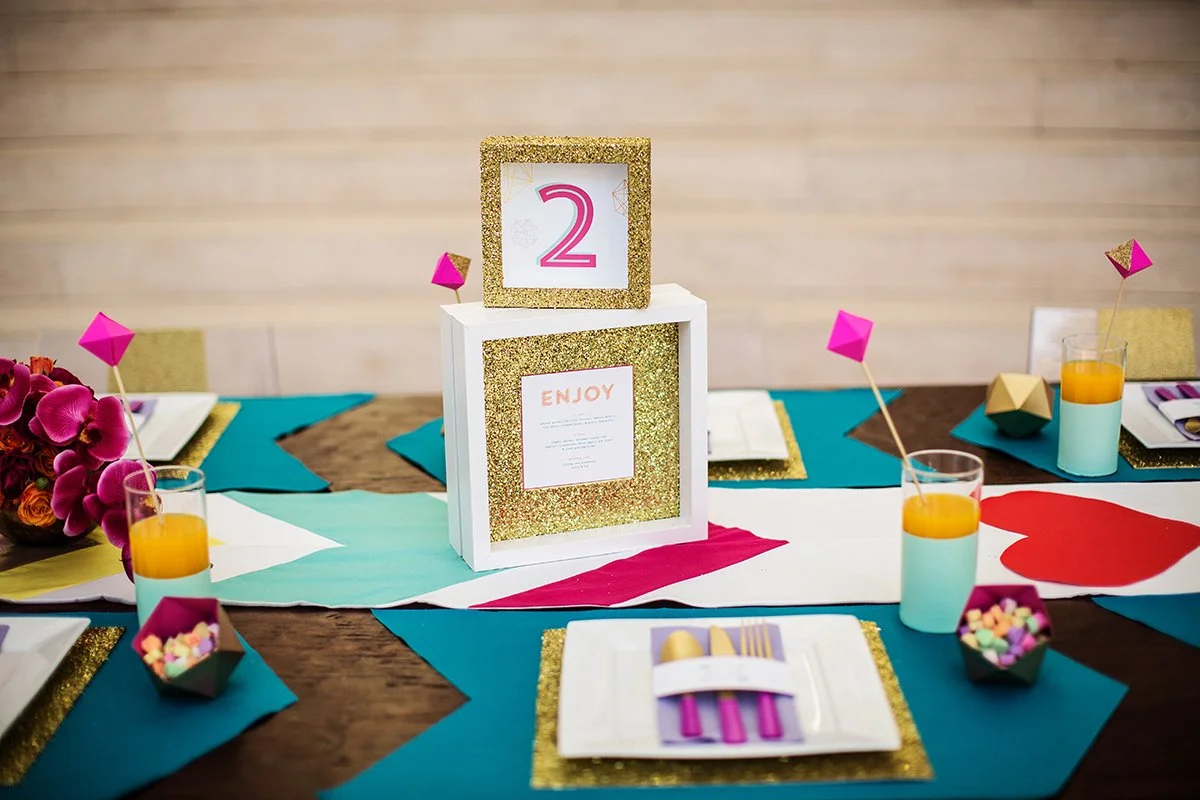 Decorative table setting for a celebration with a colorful theme. The centerpiece includes a framed number 2 and a note card with the word 'ENJOY' in gold glitter. The table has teal, white, gold, pink, and brown paper accents, with pink flower arran
