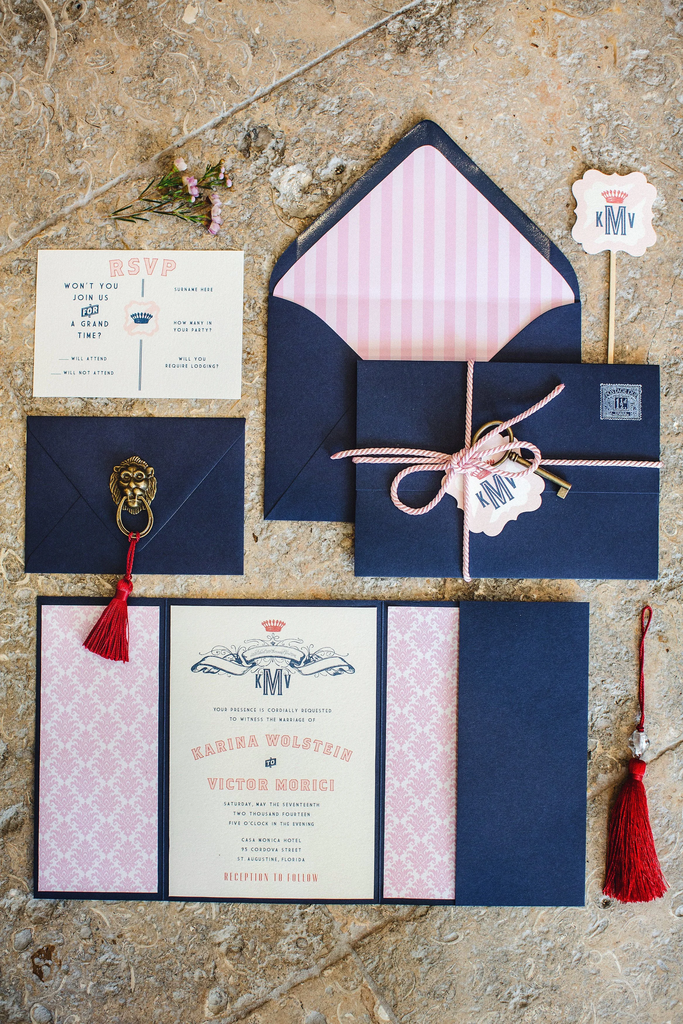 Wedding invitation set with navy blue envelopes, pink patterned paper, RSVP card, monogram sticker, keys, and decorative tassels on a stone surface.