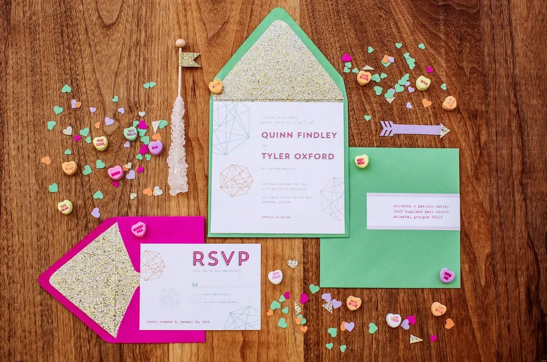 Wedding invitation suite with RSVP card on pink card stock, main invitation on white paper with geometric designs, and green outer envelope. Decorated with heart-shaped and round confetti, small candy hearts, and arrow-shaped confetti on a wooden sur