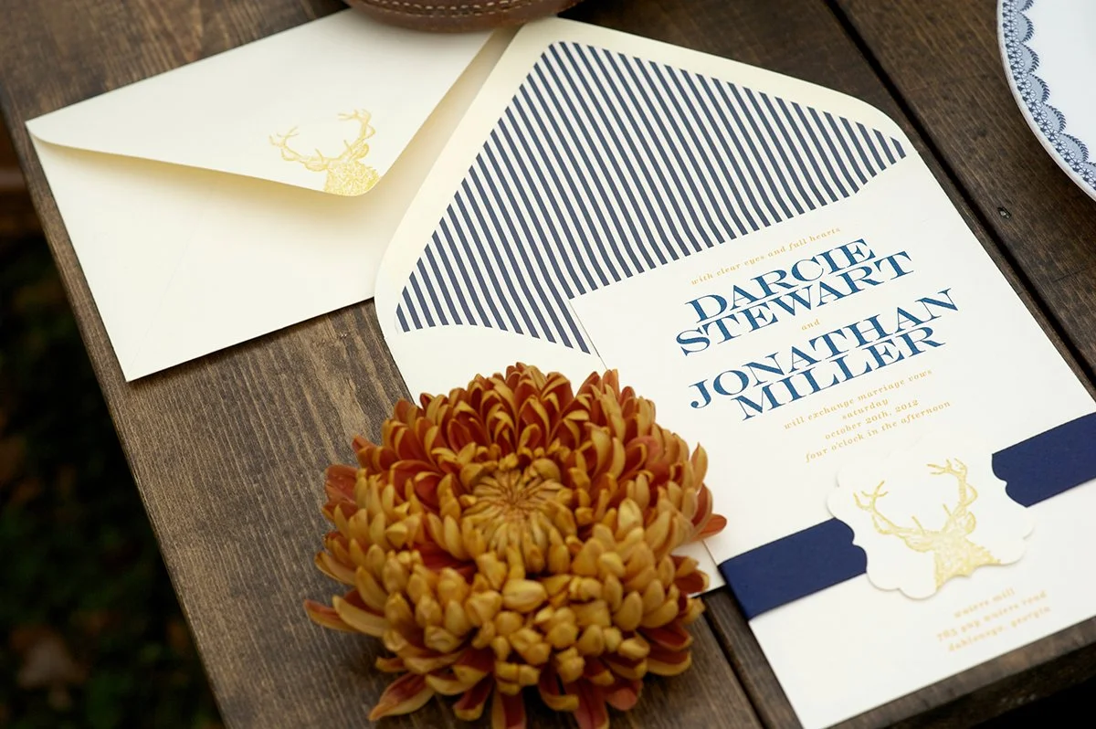 Wedding invitation featuring navy blue and gold accents, with names Darcie Stewart and Jonathan Miller, and gold-embossed deer head logo. The invitation is laid on a wooden surface next to a matching envelope with a gold deer logo and an orange flowe