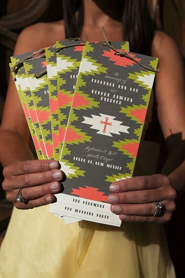 Person holding wedding programs with colorful geometric patterns, a cross symbol, and text for a wedding ceremony and reception.
