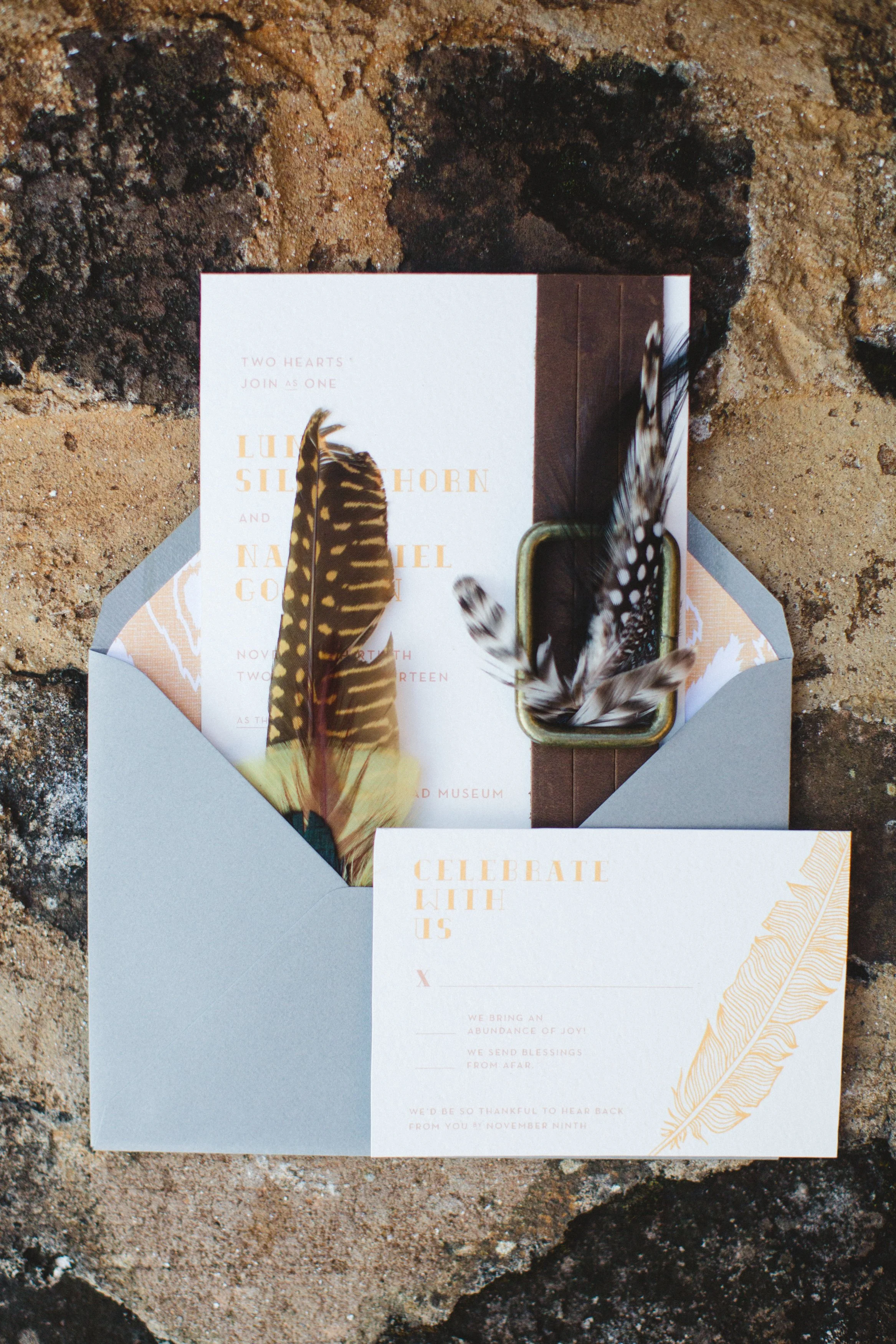 An invitation set on sandy ground with a light blue envelope, featuring feathers and text about a wedding celebration.