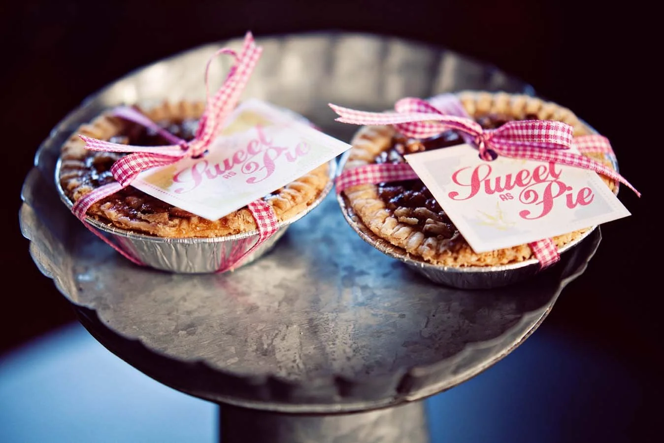 Two mini apple pies with lattice crust, tied with pink and white checkered ribbons, placed on a silver pedestal tray, each with a "Sweet as Pie" tag.