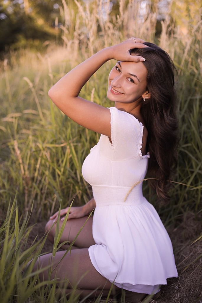 A smiling woman sits in a field of tall grass, wearing a white dress, with one hand on her head and the other on the ground, during what appears to be golden hour.
