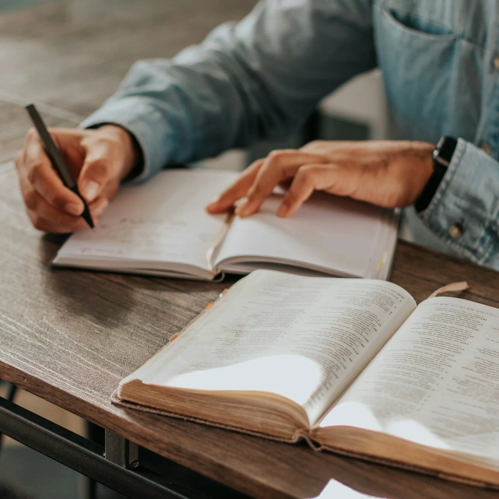Open Bible on a wooden table with text pages visible, nearby a notebook with handwritten notes and a person writing.