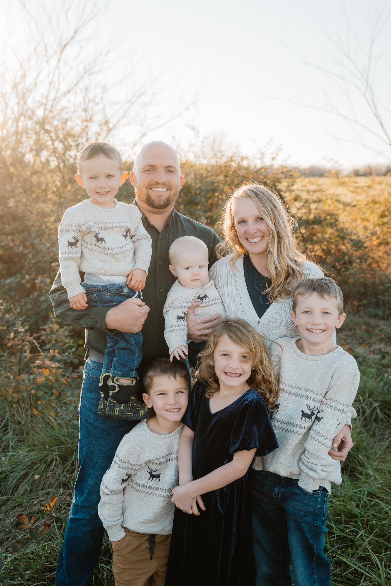A family of nine people outdoors during fall, with autumn trees in the background, smiling and dressed in cozy sweaters and casual clothing.